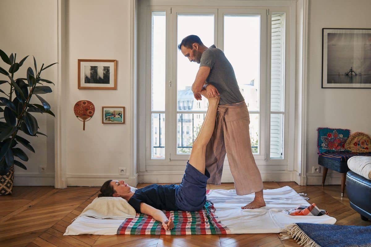 Nicolas Luc Levé standing and gently lifting the legs of a person lying on a mat, Thai style